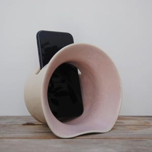 Puede incluir: Un soporte para teléfono de cerámica de color crema con interior rosa. El soporte tiene un diseño curvo único con una gran abertura. Un teléfono inteligente negro se coloca dentro del soporte, que se encuentra sobre una superficie de madera.