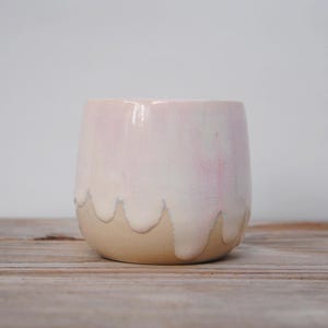 Puede incluir: Una taza de cerámica de forma redondeada. La mitad superior es de color rosa pálido, que se funde en una base beige. La pintura rosa gotea en un patrón ondulado. La taza está sobre una superficie de madera.