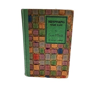 May include: A vintage book titled "Dressmaking Made Easy" by Laura I. Baldt. The cover features a colorful, textured pattern of squares in shades of orange, purple, blue, and green. The spine is green, and the book is from The McCall Company.