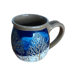 May include: A ceramic mug with a rounded body and a handle. The mug features a dark blue background with a winter scene of bare trees and a fence in gold and white. The rim and handle are a tan colour.