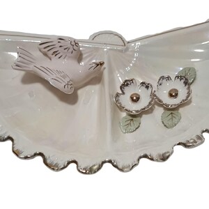 May include: A white ceramic serving dish shaped like a fan with a gold trim. The dish has two sections and features a pink bird and two white flowers with gold centers.