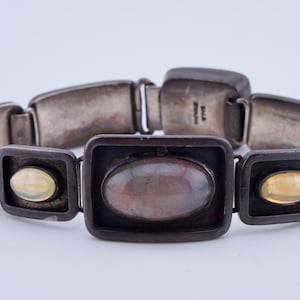 May include: A silver-toned bracelet with rectangular links and various inset stones. The bracelet features a central oval stone, flanked by smaller oval and round stones in shades of red and yellow.