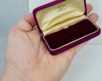 May include: A small, burgundy velvet jewelry box with a gold satin lining. The box is open and the inside is empty. The box has the inscription "E. Kaufhold Jeweler Columbia, PA" on the inside lid.