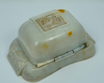 May include: A white plastic ring box with a hinged lid. The lid has a decorative design with a gold-colored pattern. The box has a worn, vintage look.
