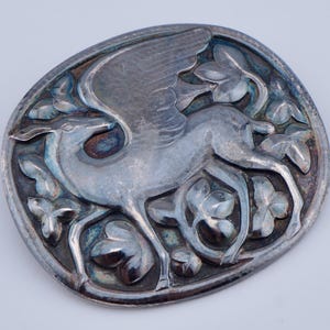 May include: An oval silver-toned metal brooch featuring a raised relief design of a winged deer surrounded by stylized leaves and floral elements. The metal surface displays a polished finish with darkened recessed areas to highlight the intricate details.