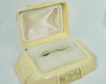 May include: A vintage, off-white ring box with a velvet interior and ornate detailing. The box is open and the lid is propped up.