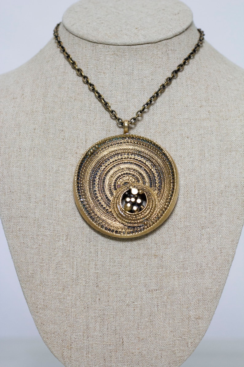 May include: A gold necklace with a large, round pendant. The pendant is made of concentric circles with a smaller circle in the center. The smaller circle has a pattern of small dots.