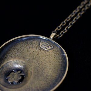 May include: A round, brass pendant with a textured surface and a smaller, circular cutout in the center. The pendant is attached to a chain necklace. The back of the pendant has the inscription "HANDMADE IN ITALY".