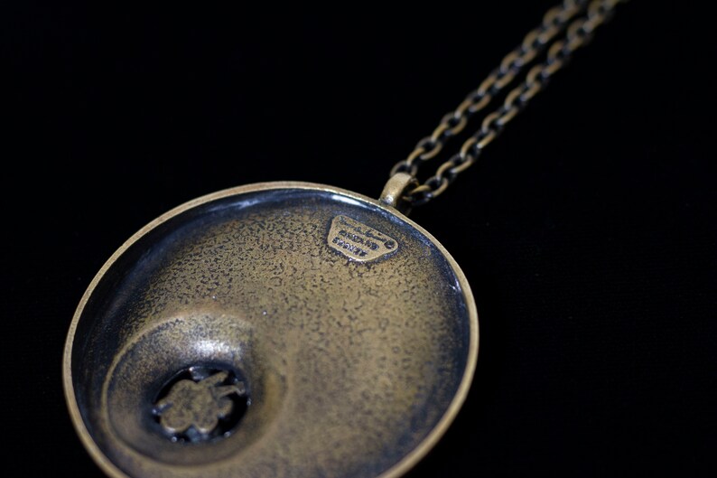 May include: A round, brass pendant with a textured surface and a smaller, circular cutout in the center. The pendant is attached to a chain necklace. The back of the pendant has the inscription "HANDMADE IN ITALY".