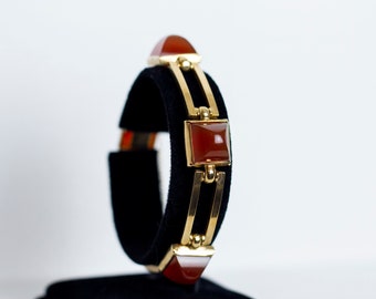 May include: A gold bracelet with a geometric design featuring square and triangular carnelian stones.
