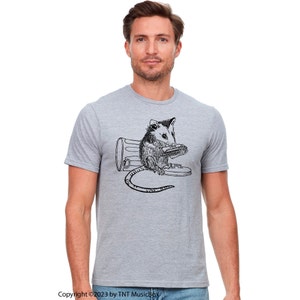 May include: A grey t-shirt featuring a black and white illustration of an opossum playing a harmonica whilst sitting in a bin.