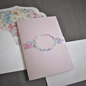 May include: A pink card with a floral design and a white envelope. The card has a scalloped edge and a floral pattern on the front. The envelope is plain white.