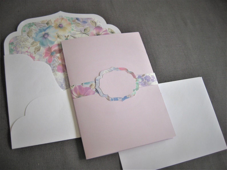May include: A pink card with a floral design and a white envelope. The card has a scalloped edge and a floral pattern on the front. The envelope is plain white.