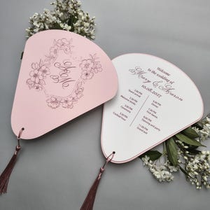 Op de afbeelding: Twee waaiervormige trouwprogramma's van lichtroze karton met donkerroze kwastjes en witte bloemen. De ene kant heeft een bloemenkader met de initialen M en A, de andere kant toont het dagschema voor de bruiloft van Mary en Aaron op 10.08.2027.