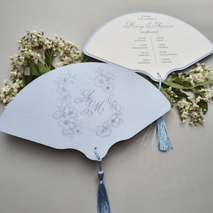 May include: Two light blue fan-shaped wedding programs with a light blue tassel. One fan features a floral frame with the monogram AM, while the other displays the text Welcome to the wedding of Mary & Aaron 10.08.2027 and a schedule of events.