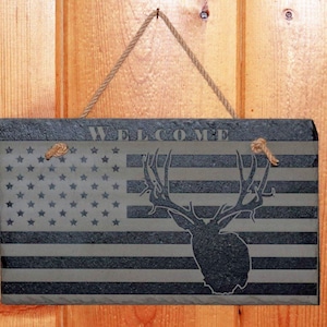May include: A rectangular welcome sign with a rustic design. The sign features the American flag with a deer silhouette. The word "WELCOME" is at the top. It hangs from a jute rope. The sign is mounted on a wooden background.