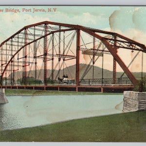 May include: Vintage postcard featuring the Delaware River Bridge in Port Jervis, N.Y. The bridge is a reddish-brown steel structure with an arched design, spanning a river. The postcard has a faded, aged appearance, with a light blue sky and green grass.