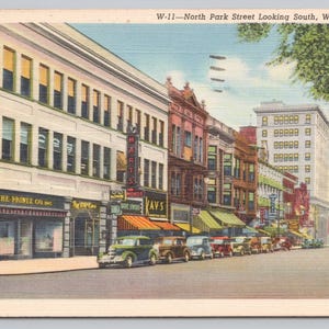 May include: A vintage postcard depicting a street scene in Warren, Ohio. The postcard shows a row of brick buildings with storefronts, including "The Printz Co. Inc." and "Kay's". Cars are parked along the street, and a tall building is visible in the background.