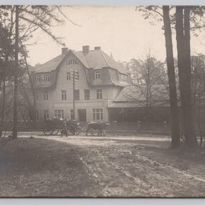 May include: A black and white vintage photograph of a large house with a horse-drawn carriage in front of it. The house has a large porch and a steep roof. The carriage is being driven by a person who is not visible in the photo.