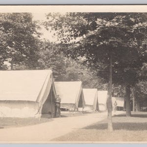 May include: A sepia-toned photograph of a military camp, featuring a row of canvas tents along a path. Trees are in the background, and a person stands near a tent. The image is a vintage postcard.