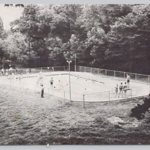 May include: A vintage black and white photograph of an outdoor swimming pool surrounded by a chain-link fence. Several people are standing near the pool, and trees are visible in the background. The pool has a rounded shape.