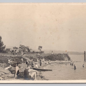 May include: A vintage black and white postcard showing a lakeside scene with people swimming and sunbathing. A man in uniform stands on the rocky shore. Other people are in the water and on the shore. A canoe is on the beach.
