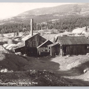 May include: Black and white vintage postcard depicting the ruins of the Matchless Mine. The image shows several weathered wooden buildings, a tall chimney, and a mountainous landscape in the background. The text "RUINS OF THE FAMOUS (TABOR) MATCHLESS MINE" is visible.