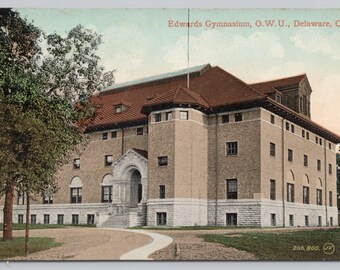 May include: A vintage postcard depicting Edwards Gymnasium at Ohio Wesleyan University in Delaware, Ohio. The building is a large, brick structure with a red tile roof and a tall, central tower. There is a large tree in the foreground.