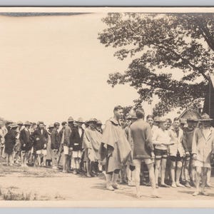 May include: A vintage black and white photograph showing a line of people outdoors. Most are wearing hats and coats. A tree and a tent are visible in the background of the image.