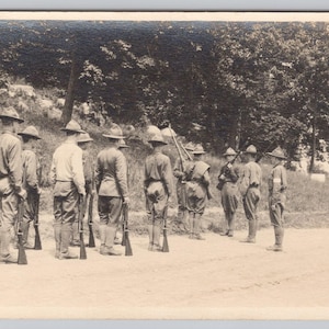 May include: A vintage black and white photograph showing a group of soldiers in uniform, standing in formation outdoors. They are wearing hats and holding rifles. The background includes trees and a dirt track.