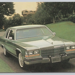 May include: A vintage green Cadillac sedan driving on a curved road. The car has a chrome grille, white-walled tires, and a license plate. The background features trees, grass, and a building.