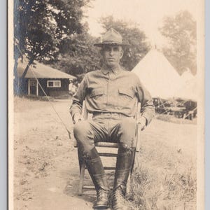 May include: A sepia-toned photograph depicting a man in military attire seated on a wooden chair. He is wearing a hat, shirt, trousers, and boots. The background includes a small building and tents, suggesting a camp setting.