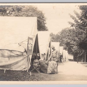 May include: A monochrome photograph depicting a military camp. Canvas tents are arranged along a path, with soldiers and equipment present. The image conveys a sense of history and military life from a bygone era.