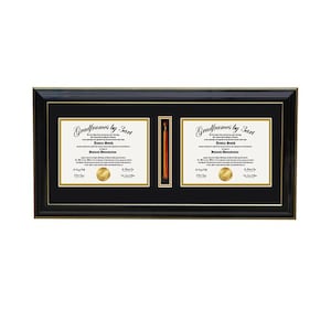 May include: A black rectangular diploma frame with gold trim, featuring two white document slots and a central vertical compartment containing an orange tassel. The frame includes black matting with gold accents around each document and the tassel.