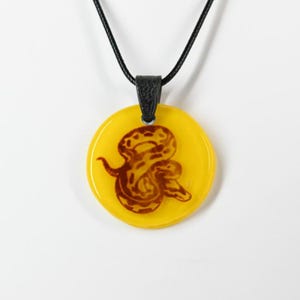 May include: A circular pendant featuring a brown snake illustration encased in a bright yellow translucent material. The pendant is attached to a black cord by a textured black bail, creating a contrast between the vibrant yellow and dark elements.
