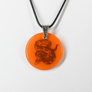 May include: A round, bright orange pendant featuring a dark brown snake silhouette in the center. The pendant is attached to a silver-toned metal bail with an etched pattern and hangs from a thin, smooth black cord necklace.