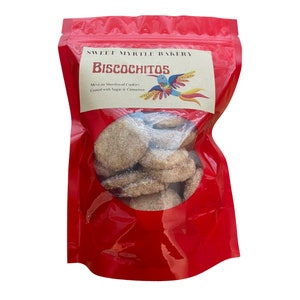 May include: A red resealable bag with a label that reads "Sweet Myrtle Bakery Biscochitos Mexican Shortbread Cookies Coated with Sugar & Cinnamon". The bag contains a pile of biscochitos, which are Mexican shortbread cookies.