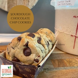 May include: Close-up of four sourdough chocolate chip cookies in a brown paper baking pan. The cookies are golden brown and have chocolate chips throughout. The text "SOURDOUGH CHOCOLATE CHIP COOKIES" is in a brown circle above the cookies. The text "GEORGIA grown" is in a green circle with a leaf design in the lower left corner of the image.