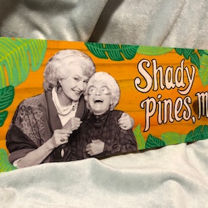 May include: A wooden sign with a yellow background and green tropical leaves painted on the edges. The sign features a black and white photo of two women smiling and the text "Shady Pines, Ma!"
