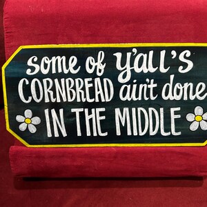 May include: A rectangular wooden sign with a black background and yellow trim. The sign features white lettering that reads "some of y'all's cornbread ain't done in the middle." Two small white flowers with yellow centers are on the sign.