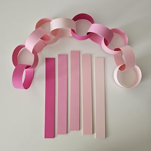 May include: A paper chain in shades of pink and a set of pink paper strips. The chain is made of connected loops, with colors ranging from light to dark pink. The strips are arranged vertically, showcasing a gradient of pink hues.