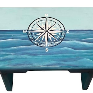 Op de afbeelding: Een houten opstapje met een nautisch thema. De bovenkant is verdeeld in twee delen: een lichtblauwe lucht en een blauwe oceaan met witte golven. Een kompasroos staat in het midden van de lucht. Het krukje heeft twee donkerblauwe poten.