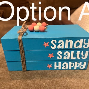 Puede incluir: Una pila de tres cajas de madera azules atadas con cuerda. Cada caja tiene las palabras "sandy", "salty" y "happy" en blanco, con una estrella de mar. La parte superior está decorada con cuentas de madera.