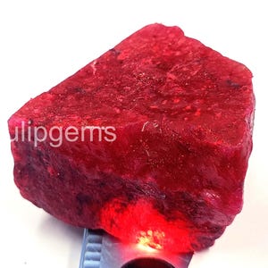 Könnte beinhalten: Ein grob geschnittener, tiefroter Rubin-Edelstein mit strukturierter Oberfläche. Der Rubin wird von einer Lichtquelle beleuchtet, die seine satte Farbe und natürliche Form zeigt. Der Text "tulipgems" ist sichtbar.