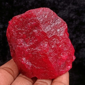 May include: A raw, uncut ruby gemstone with a deep red color. The surface of the stone has a rough, textured appearance. The gemstone is held in a hand against a dark background. This natural ruby is a mineral specimen.