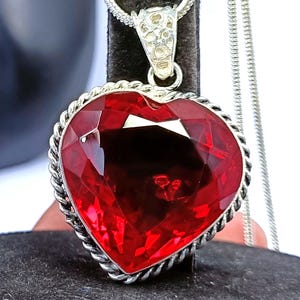 May include: A heart-shaped pendant necklace with a large, faceted red gemstone. The gemstone is set in a silver-toned metal frame with a rope-like border. The necklace has a delicate silver chain.
