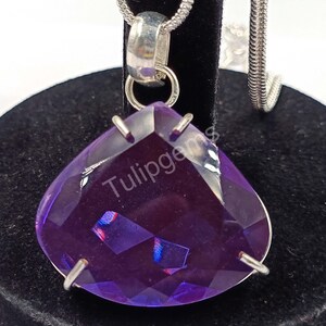 May include: A faceted, heart-shaped purple gemstone pendant with a silver chain. The pendant is a deep violet color with a polished surface and is suspended from a silver bail. The text "Tulipgems" is visible on the gemstone.