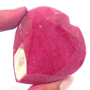 Pode incluir: Uma pedra preciosa facetada em forma de coração, em tom magenta profundo. A gema tem uma superfície polida com múltiplas facetas, que capturam a luz. Uma pequena área mais clara é visível em um lado. A gema é segurada por um dedo.