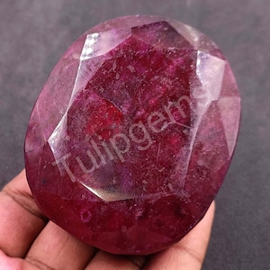 Puede incluir: Una gran gema de rubí facetada, de color rojo intenso. La piedra de forma ovalada tiene múltiples facetas que capturan la luz, creando un efecto brillante. El texto "Tulipgems" es visible en la superficie.