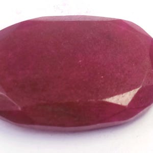 Peut inclure: Une pierre précieuse de rubis taillée en forme ovale. La gemme est d'un rouge profond et riche avec de multiples facettes qui captent la lumière. La pierre est posée sur un fond blanc.